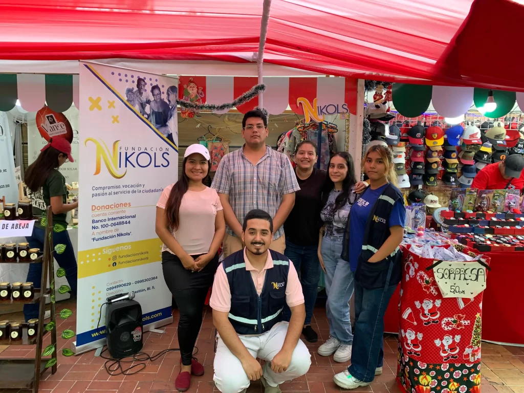 La imagen muestra al equipo de Fundación Nikols participando activamente en una feria comunitaria. En la fotografía se puede apreciar a seis personas del equipo, incluyendo voluntarios y organizadores, posando frente a un banner con el logo y la misión de la fundación. El estand de la fundación está decorado con motivos navideños. Al fondo, hay un colorido despliegue de gorras, juguetes y otros artículos, debido a que se encuentra en la feria artesanal de Malecón 2000. El ambiente es alegre y dinámico